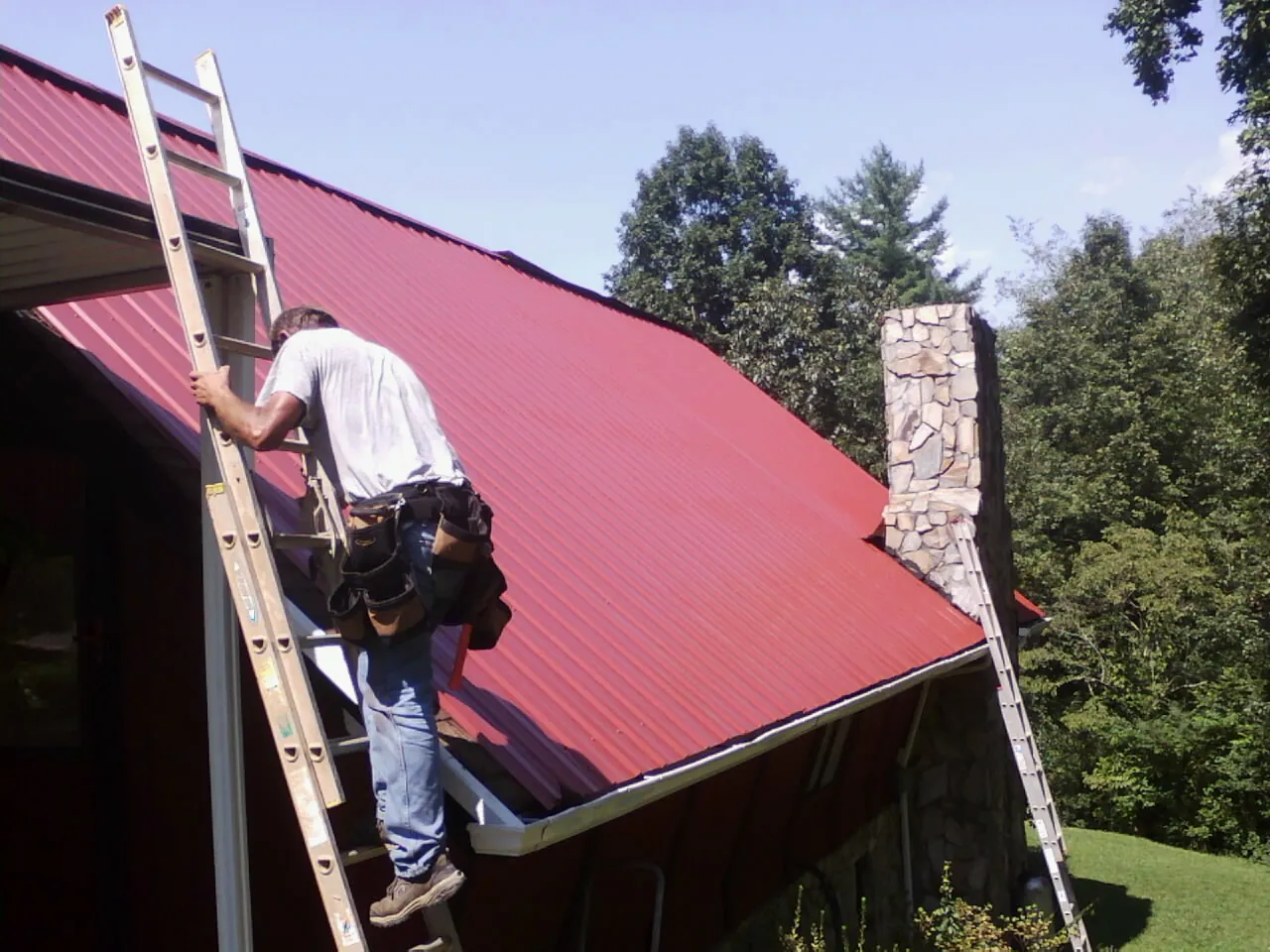 Roofer on a ladder beside a red metal roof and stone chimney — Soffit & Fascia Repair by Roofing Brothers in Sartell